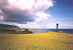 Two ahu at Hanga Roa. In foreground Ahu Ko Te Riku (with a pukao on its head). In the mid-ground is a side view of an ahu with five moai showing retaining wall, platform, ramp and pavement. The Mataveri end of Hanga Roa is visible in the background with Rano Kau rising above it.