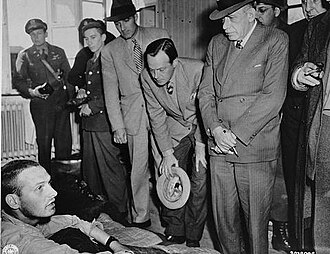 American newspaper editors speak with survivors at a hospital in the newly liberated Buchenwald concentration camp, April 1945. Left to right: Norman Chandler, Los Angeles Times; William I. Nichols (leaning forward, center), This Week magazine; and Julius Ochs Adler, The New York Times.