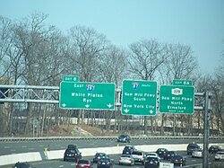 A multilane freeway with a display of three green signs over the road. The left one reads exit 8 east Interstate 287 White Plains Rye three downward arrows, the middle one reads Interstate 87 south Saw Mill Parkway south New York City two downward arrows, and the right one reads exit 8A New York State Route 119 Saw Mill Parkway north Elmsford right lane.